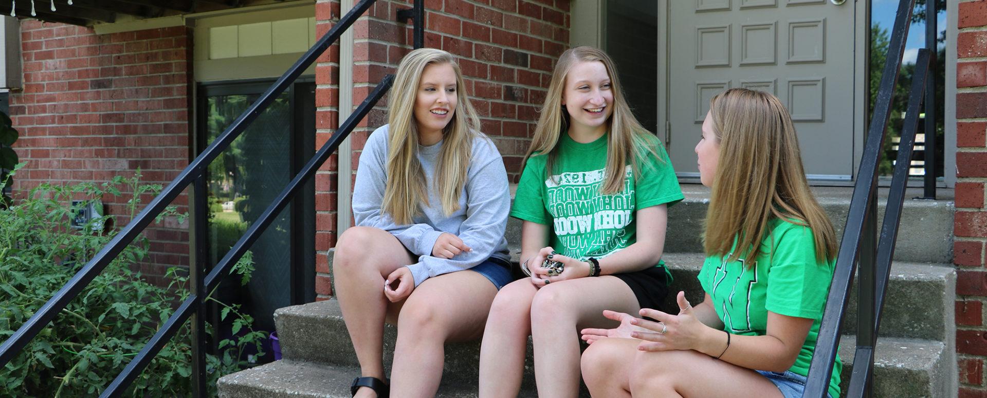Three students sit on stairs in front of an apartment
